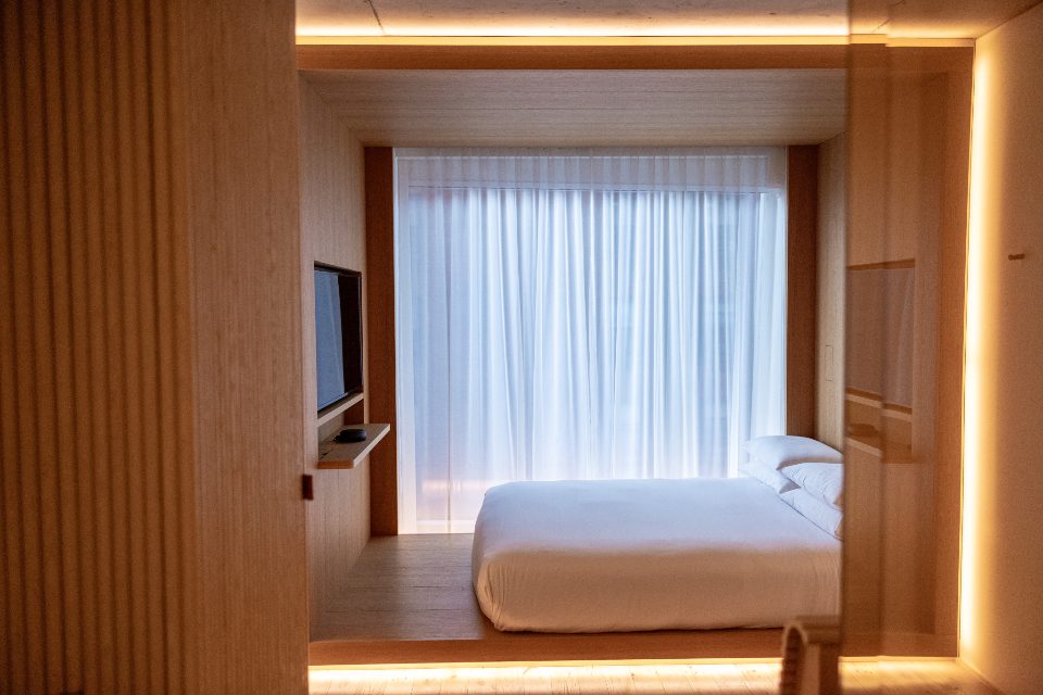 A minimal hotel bedroom in warm wood, with a low bed framed by a lit alcove and a curtained window.