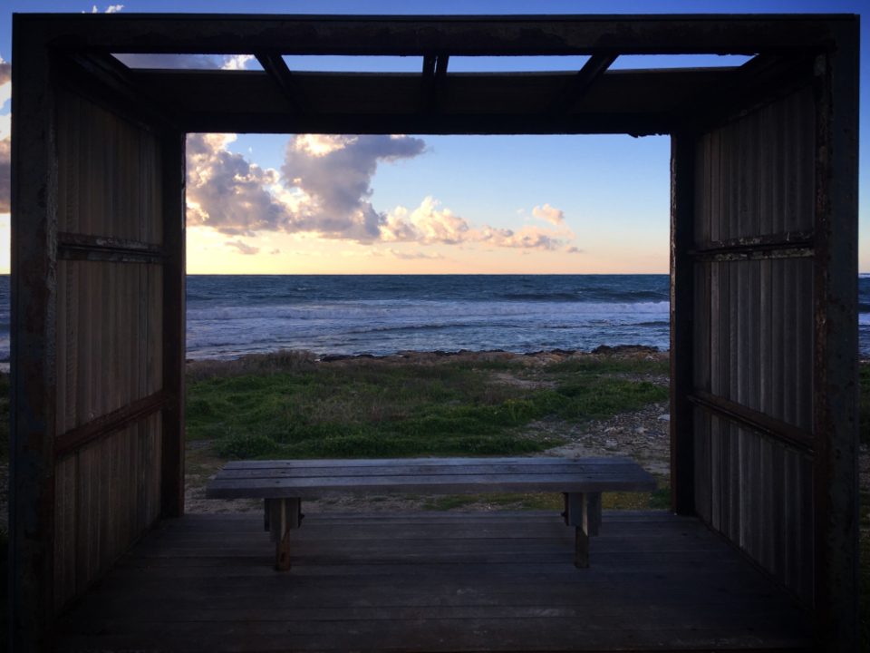 A darkened room opening onto a wide bright view of water and sky through a full-height opening.